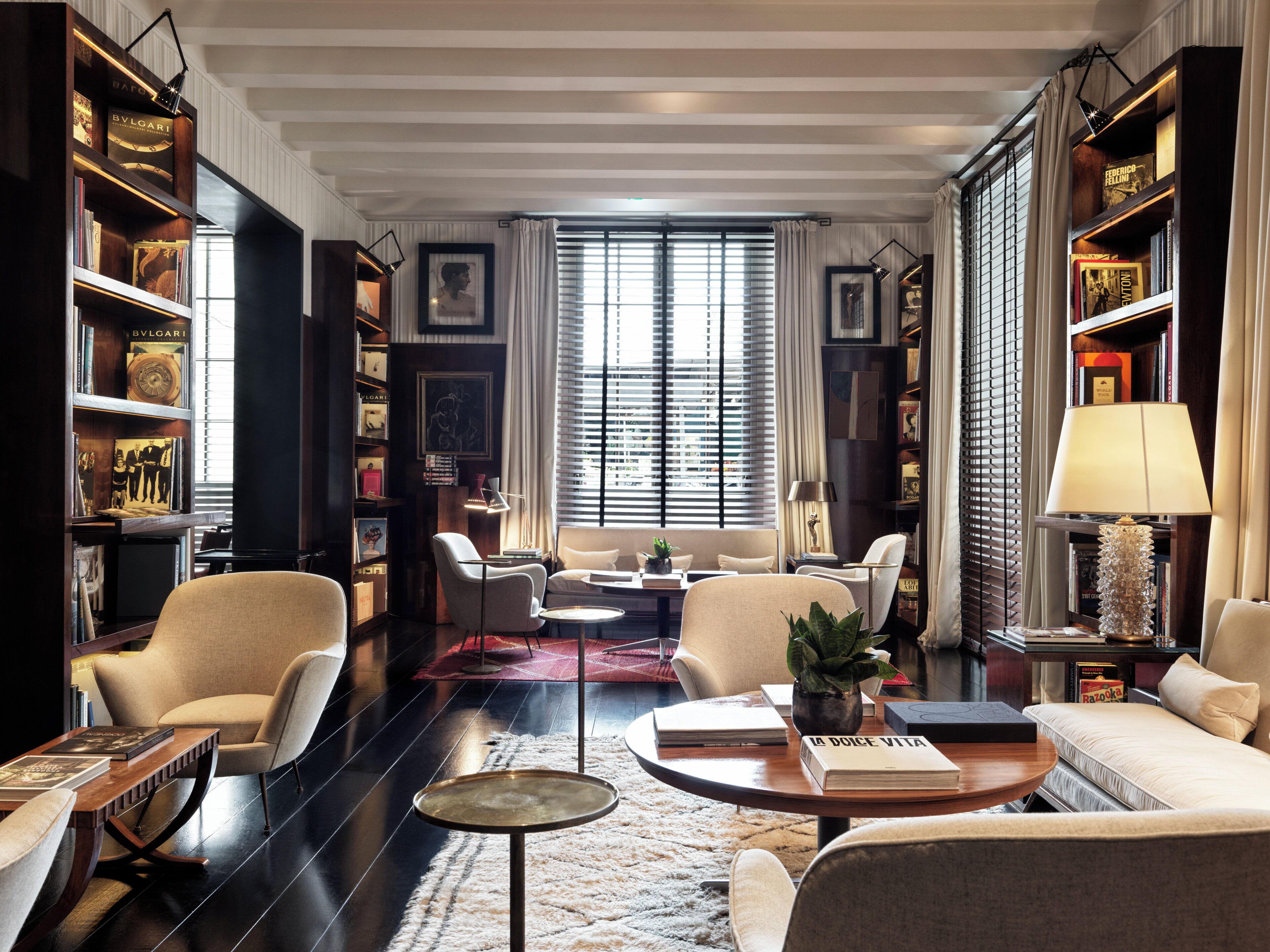 A spacious living room with a large window, multiple seating areas, and a bookshelf filled with books and decorative items.