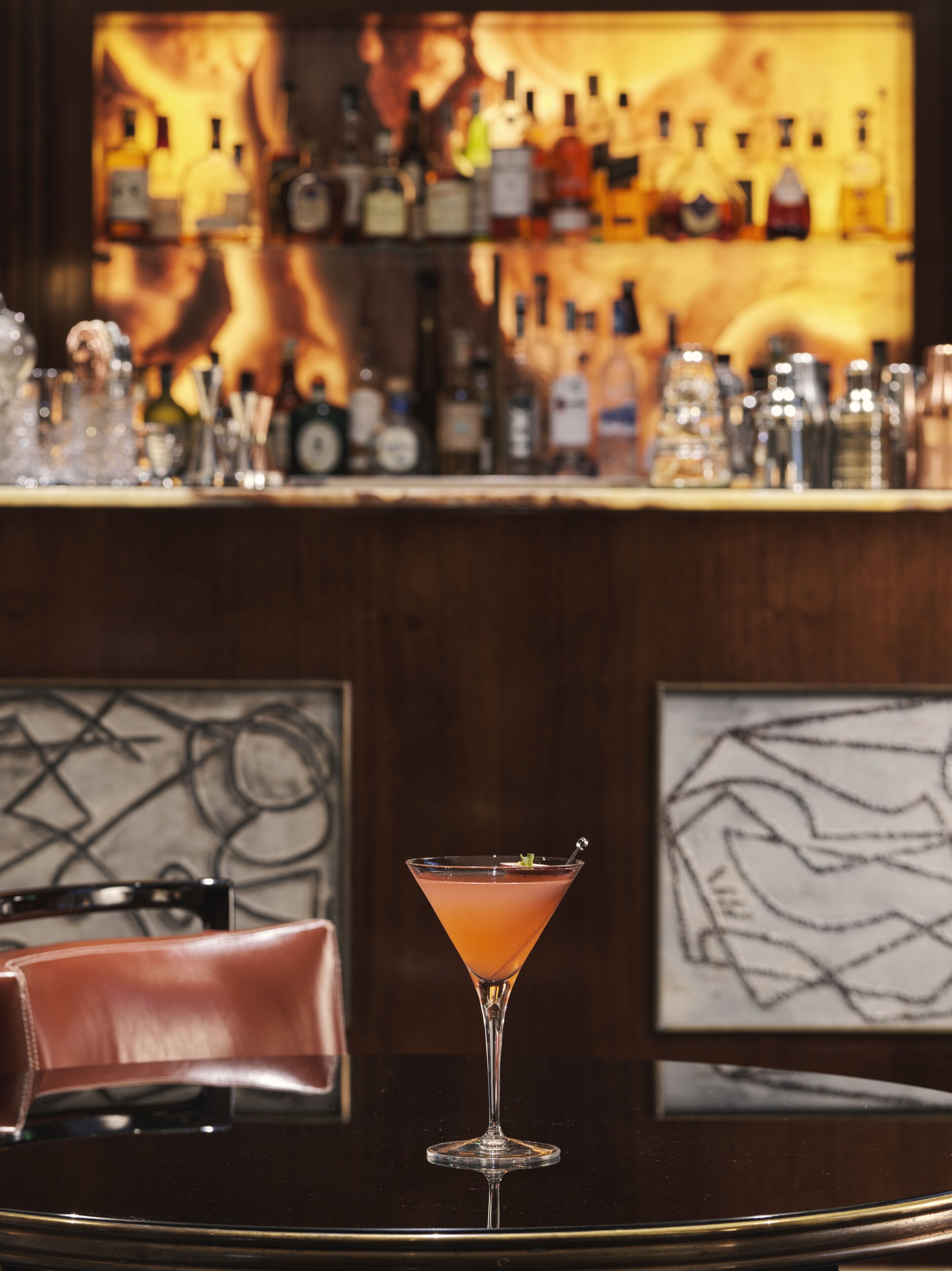 A bar area with a martini glass on a table, a bar counter with various bottles, and abstract wall art.