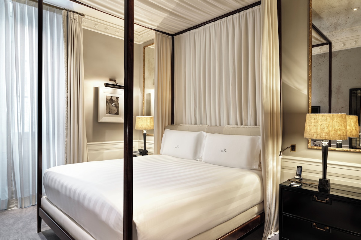A four-poster bed with a canopy, a bedside table with a lamp, and a large mirror on the wall.