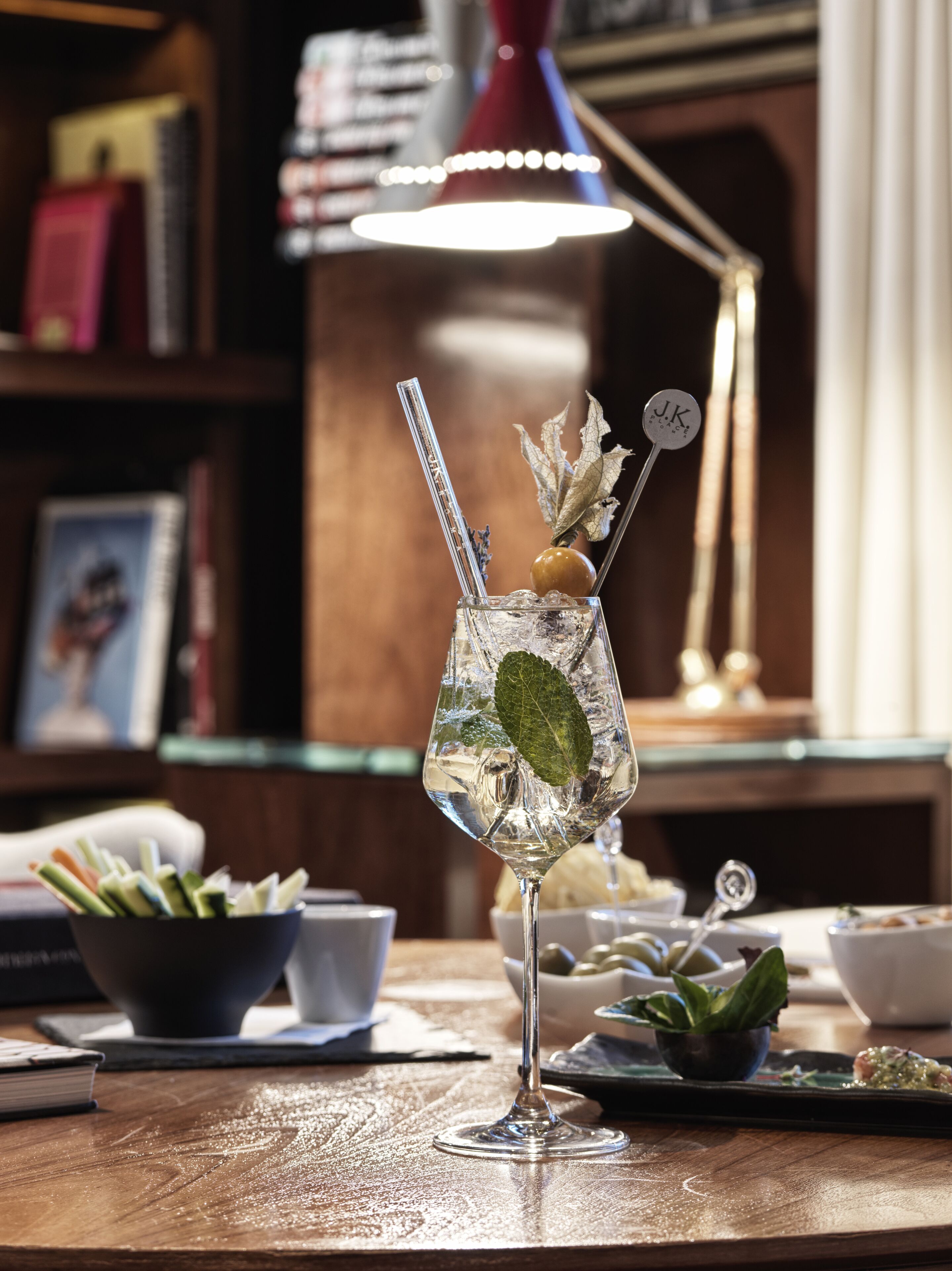 A cocktail glass with a straw and garnish on a wooden table, surrounded by various bowls and a lamp in the background.