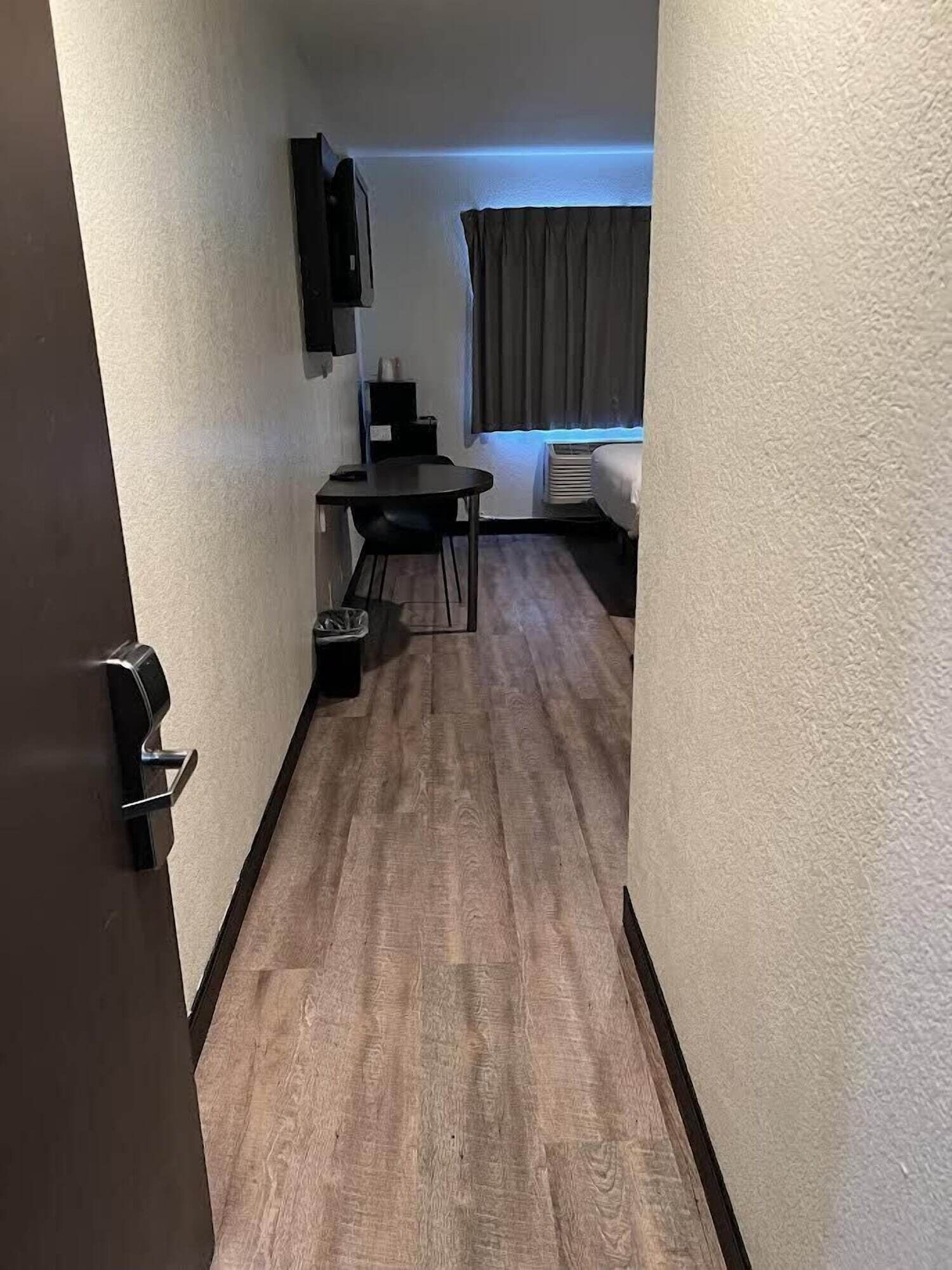 Un couloir étroit avec un plancher en bois, une petite table avec une chaise, un lit près d’une fenêtre et un téléviseur fixé au mur.