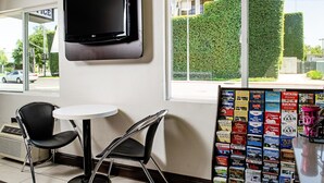 Quarto de hotel com televisão de tela plana na parede, uma mesinha redonda, duas cadeiras pretas e uma janela com vista para um sebe verde e um edifício.