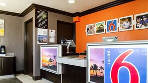 Balcão de recepção de motel com um grande sinal em formato de '6', uma mesa com um computador e uma parede com diversos cartazes e avisos.