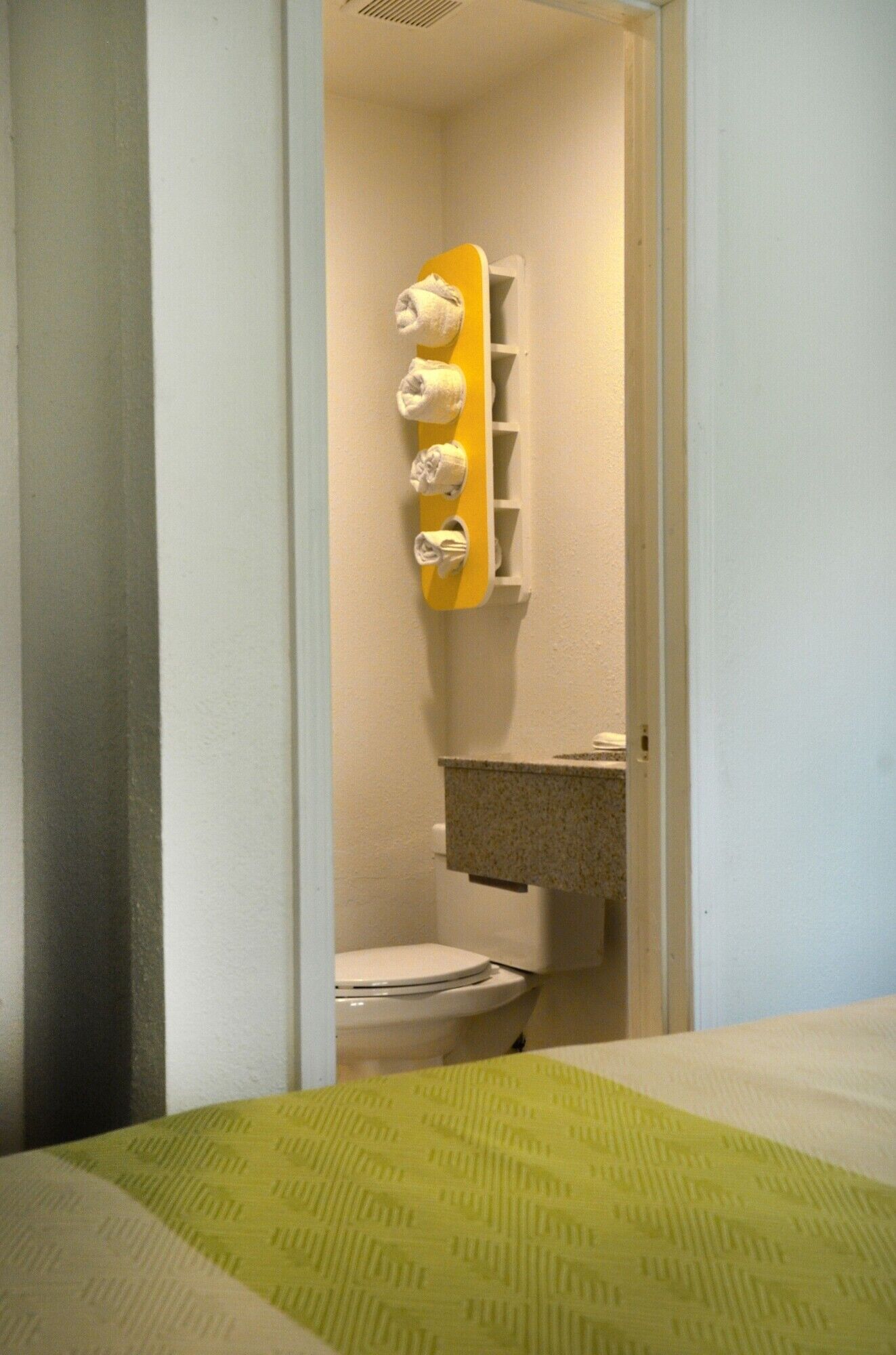 A small bathroom with a toilet, a yellow towel rack, and a green patterned bedspread.