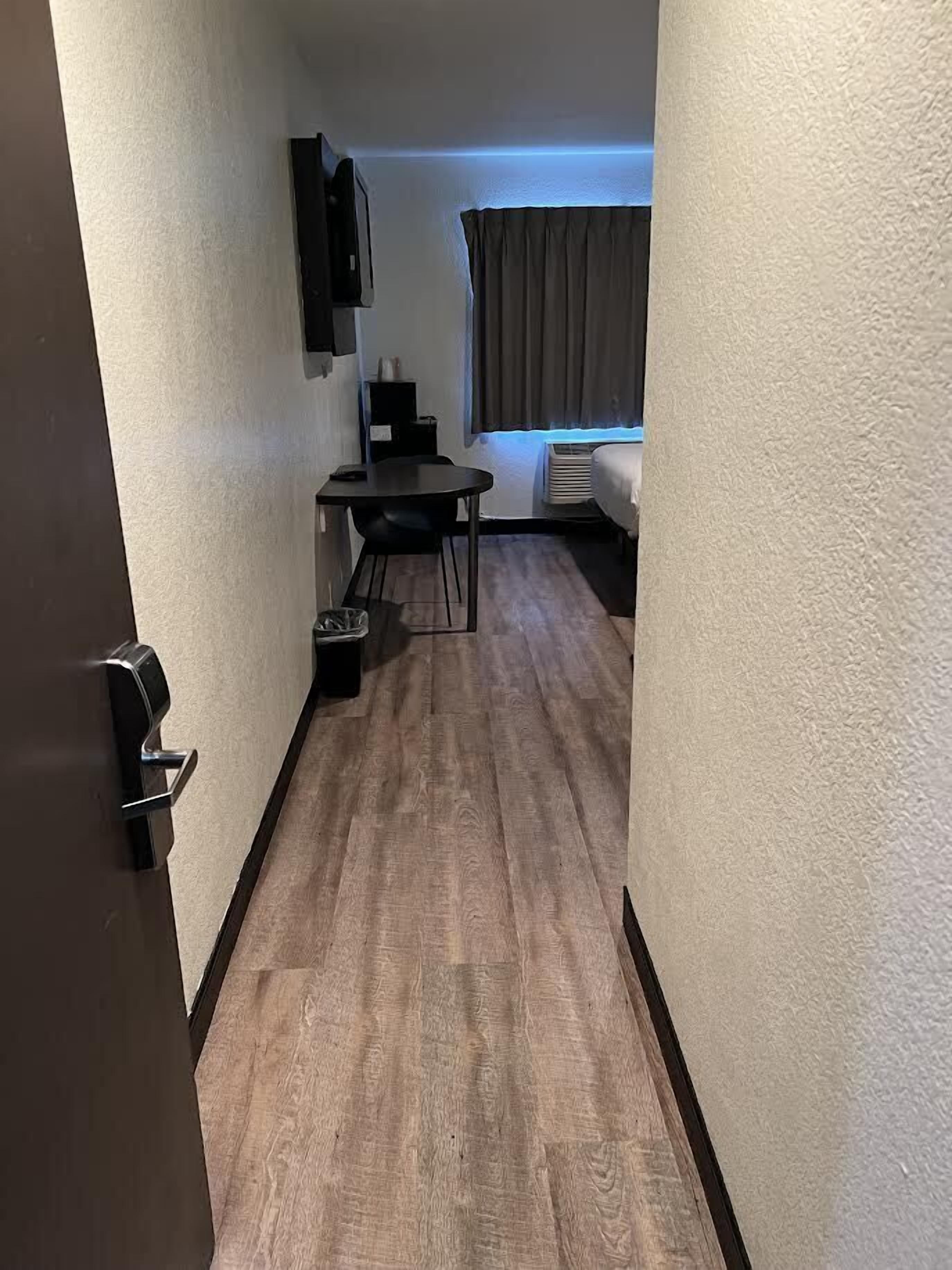 A narrow hallway with a wooden floor, a small table with chairs, a TV mounted on the wall, and a window with curtains.