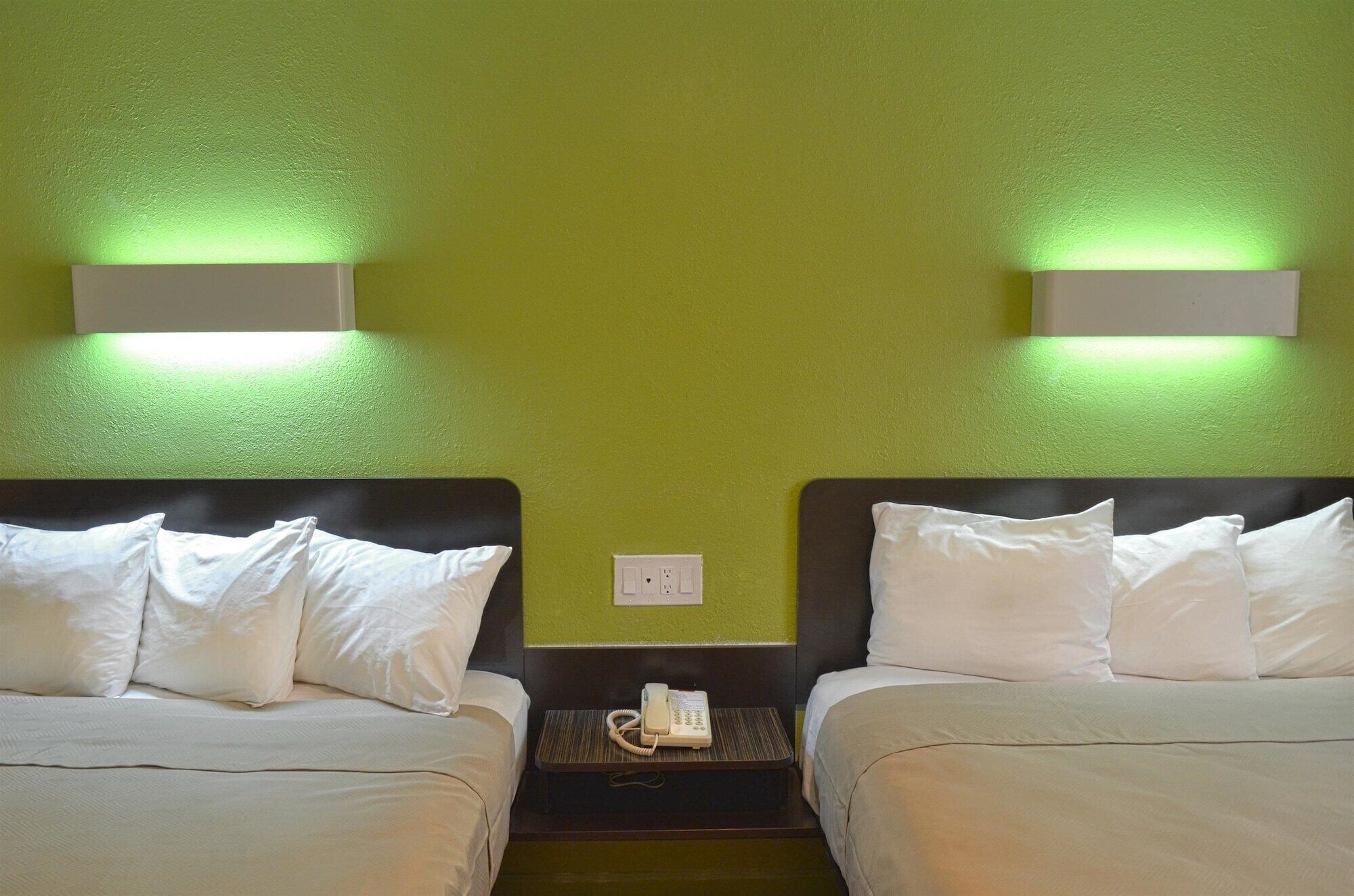 Une chambre d’hôtel avec deux lits, chacun doté de draps blancs et d’une tête de lit. Une table de chevet se trouve entre les lits, avec un téléphone et une prise électrique. Les murs sont peints en vert, et deux appliques murales sont fixées au-dessus des lits.