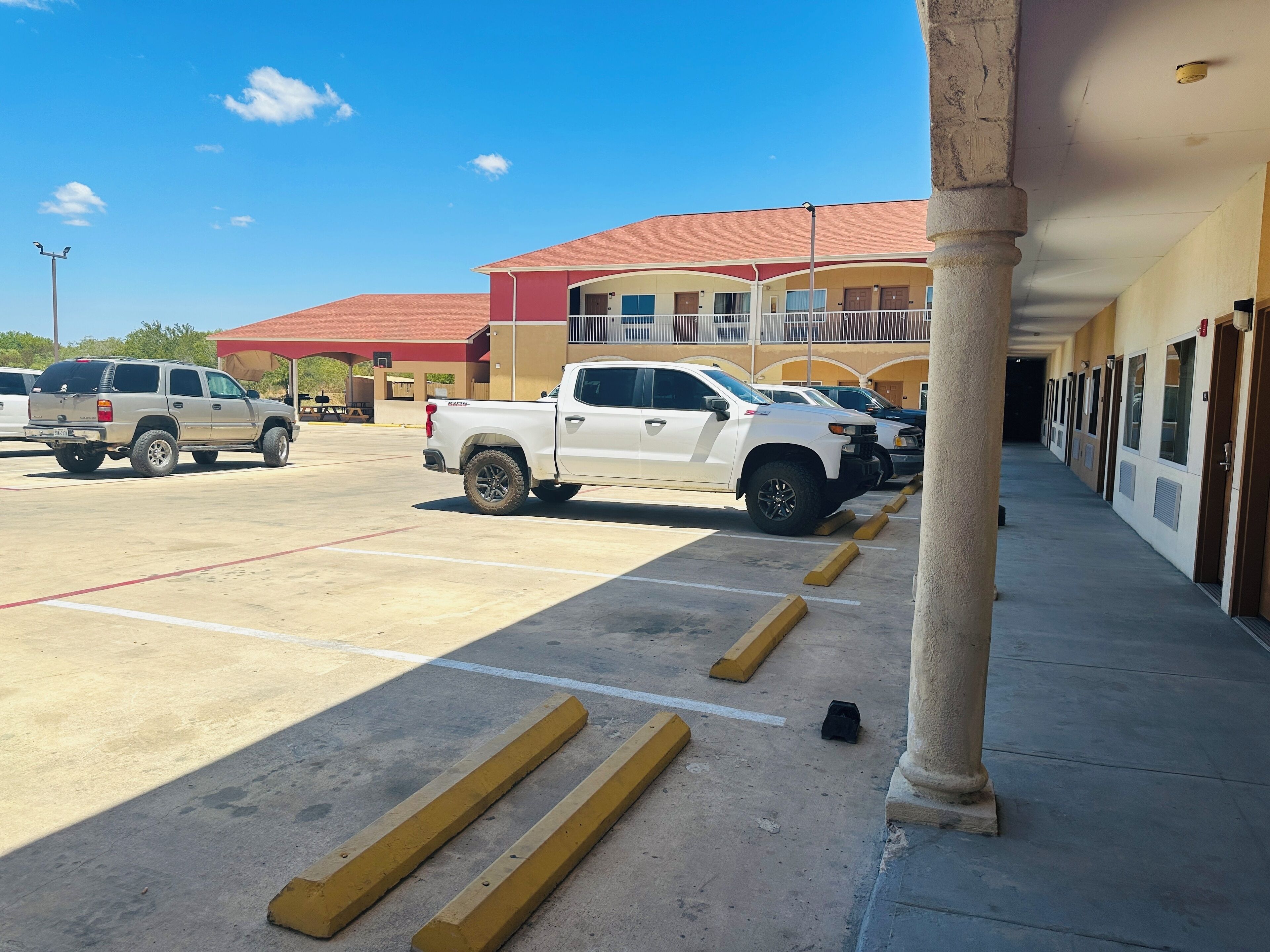 A parking lot with a white pickup truck and a beige SUV. There are yellow bollards and a building with multiple doors and windows in the background.