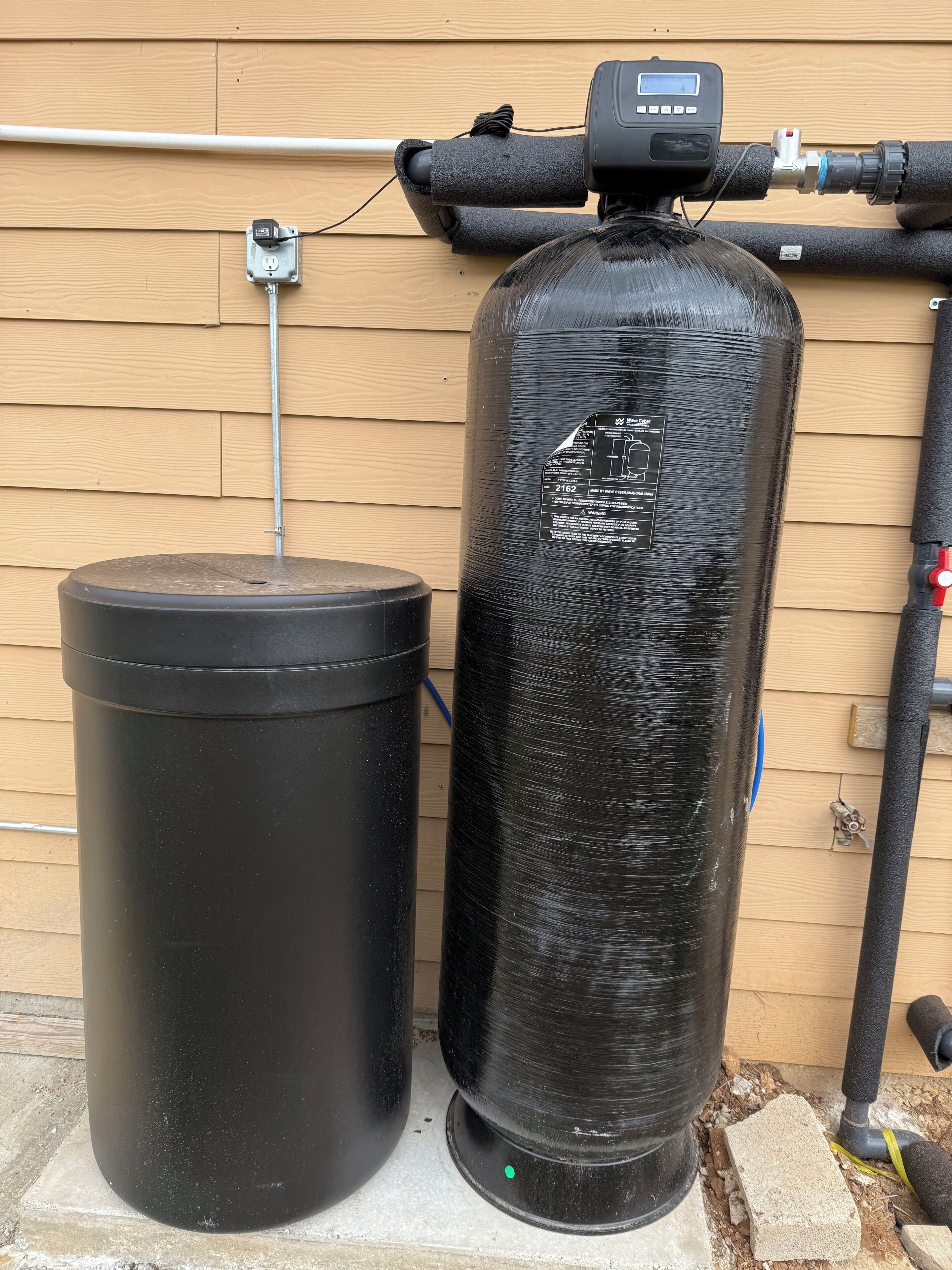 A water heater and a black trash can against a wooden wall.