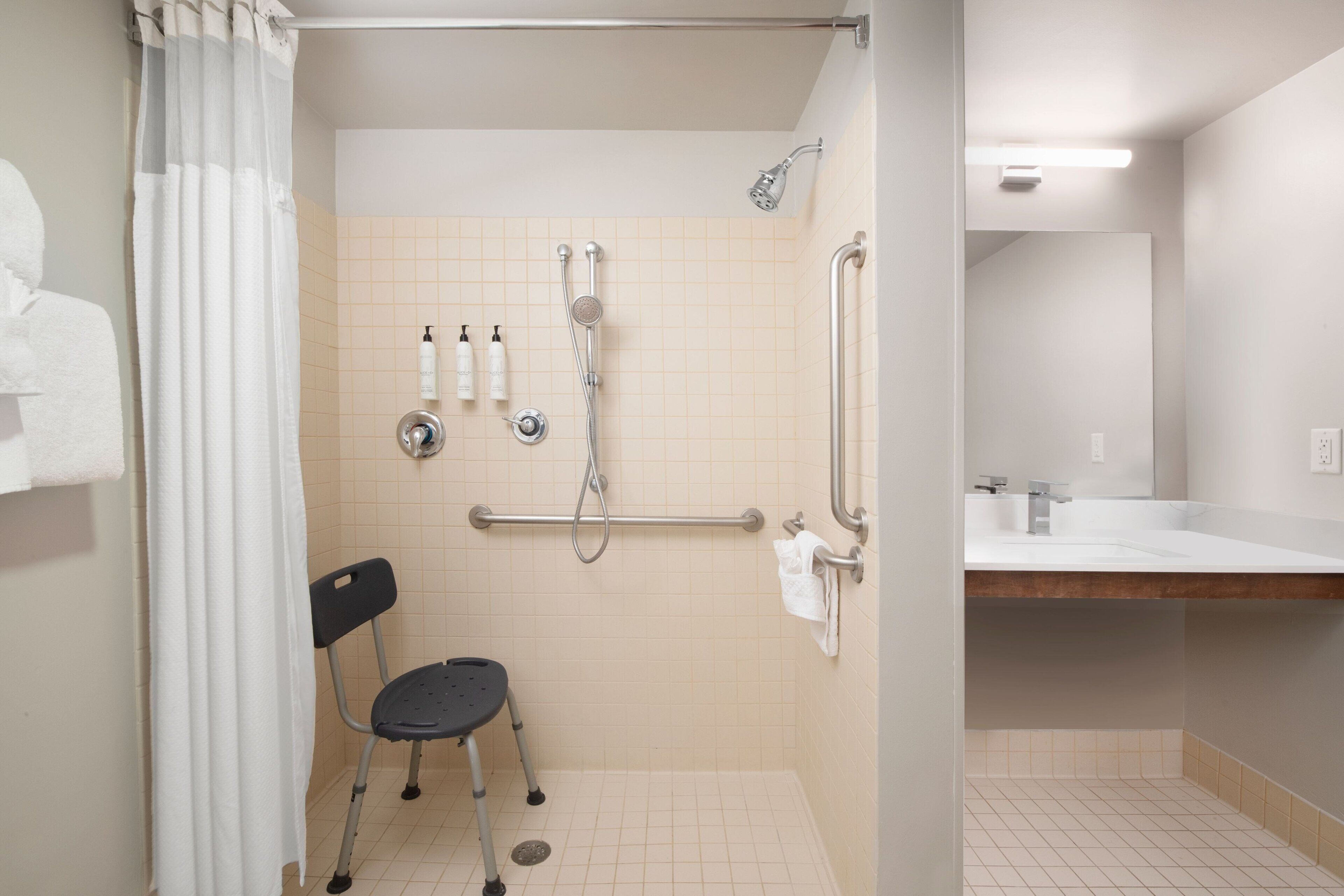 A bathroom with a shower, grab bars, a wheelchair-accessible chair, and a sink.