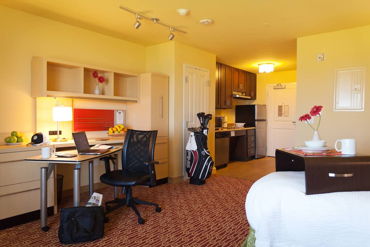 A hotel room with a bed, desk, chair, kitchenette, and a small bathroom.