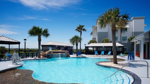 A swimming pool with a waterfall feature, surrounded by palm trees and lounge chairs. - TownePlace Suites by Marriott Galveston Island (Galveston)