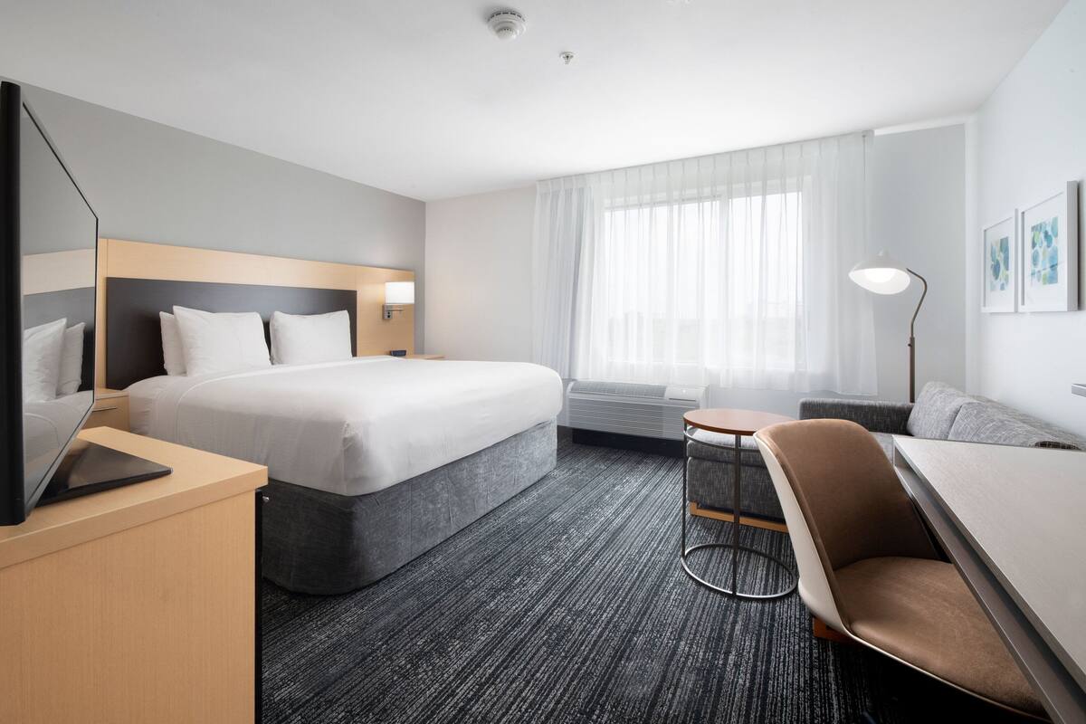 A modern hotel room with a large bed, a desk with a laptop, a sofa, and a small round table.