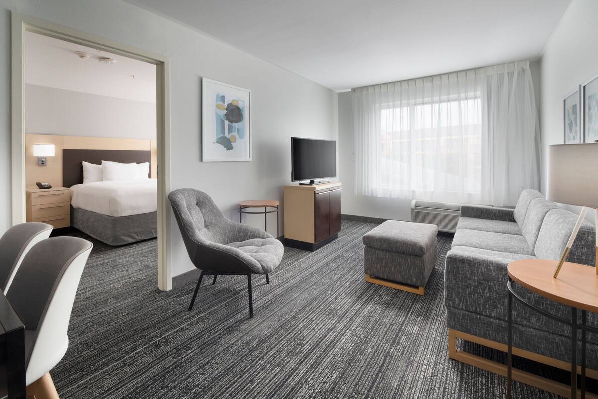 A modern hotel room with a bed, a gray armchair, a sofa, a TV, and a small table.
