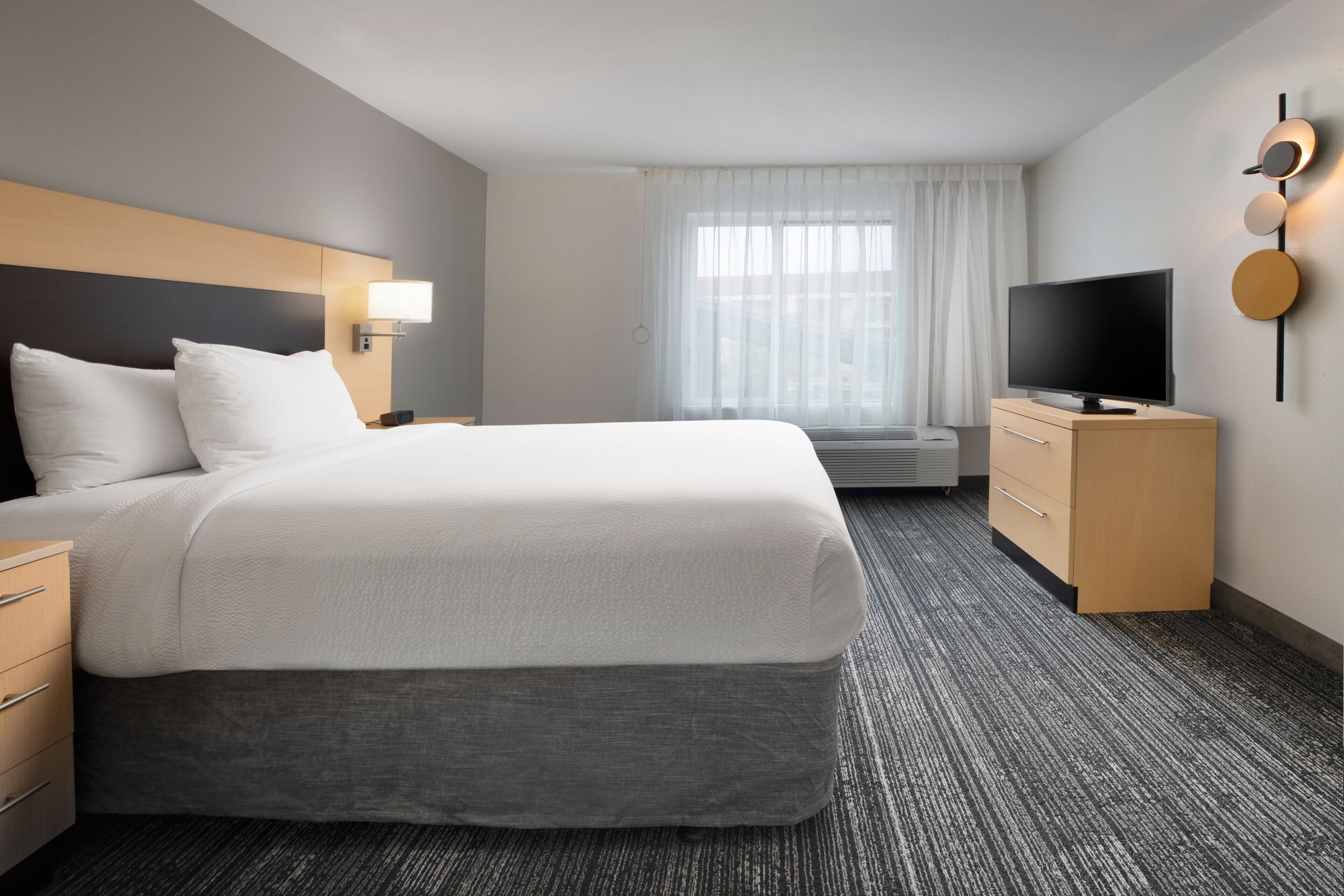 Ett modernt hotellrum med en stor säng, en platt-TV, en byrå och ett fönster med tunna, genomskinliga gardiner.