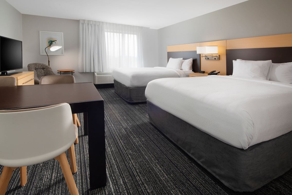 A hotel room with two beds, a desk, a chair, a TV, and a lamp.