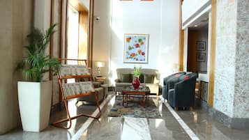 A spacious lobby with a marble floor, a seating area with sofas and armchairs, a large potted plant, and a framed artwork on the wall.