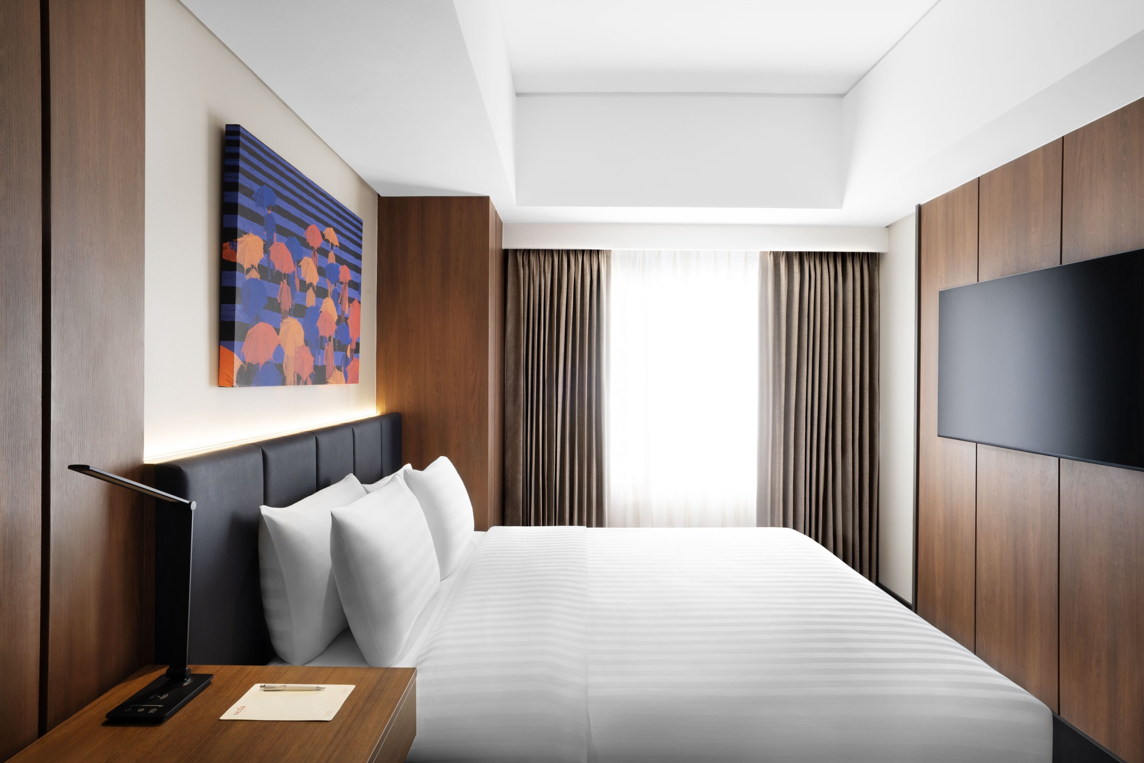 A modern hotel room with a large bed, a flat-screen TV, a desk with a lamp, and a painting on the wall.