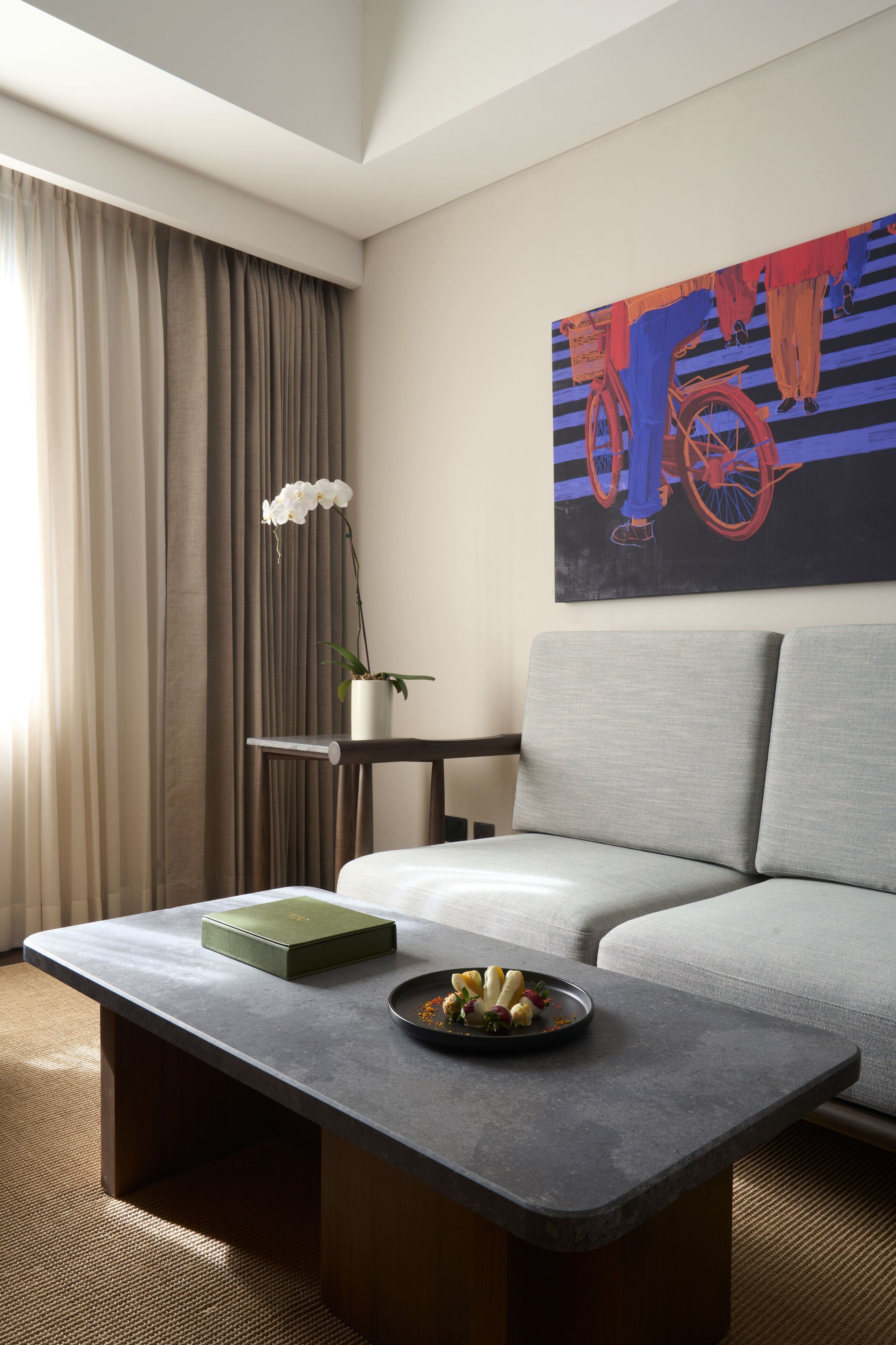 Ett modernt hotellrum med en grå soffa, ett litet runt bord med en grön låda och en tallrik med frukt, en orkidé i en kruka och en färgstark, abstrakt målning på väggen.