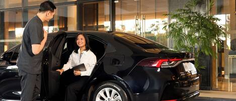 Un coche negro con una mujer sentada en su interior, un hombre de pie a su lado y una planta grande en maceta al fondo.