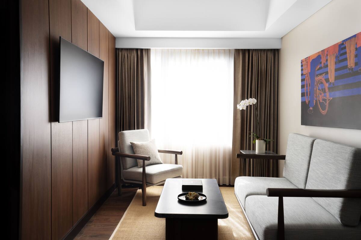 Ein modernes Hotelzimmer mit Flachbildfernseher, einer Couch, einem Sessel, einem kleinen Tisch und einem Fenster mit Vorhängen.