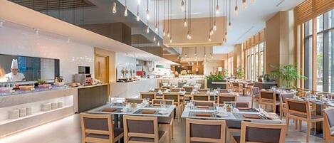 A modern restaurant with wooden tables and chairs, a kitchen area, and large windows allowing natural light.
