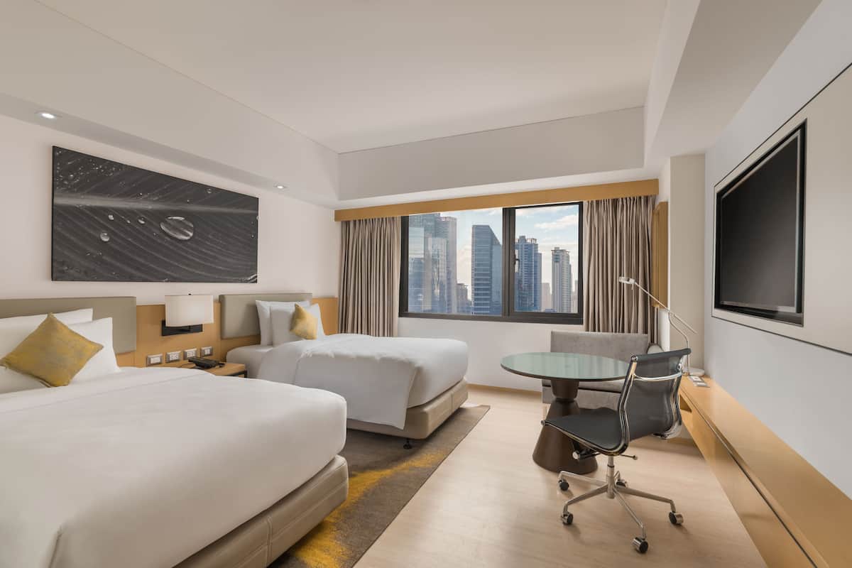 Ein Hotelzimmer mit zwei Betten, einem Schreibtisch, einem Sessel und einem Fernseher, mit Blick auf die Stadt.