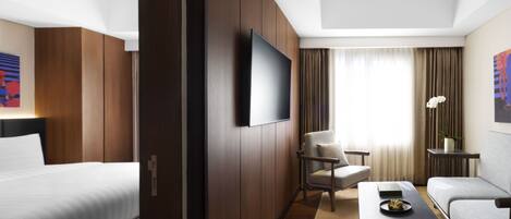 Habitación de hotel con cama, sofá, mesa de centro, televisión montada en la pared y ventana con cortinas.