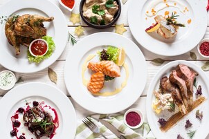 Variedad de platos en blanco, incluyendo pollo asado, salmón y chuletas de cordero, dispuestos sobre una mesa de madera.