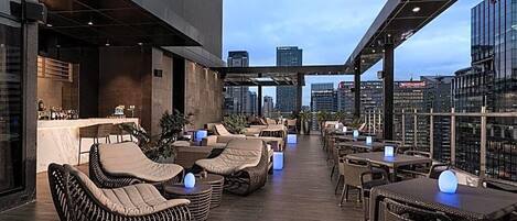 Un bar en la azotea con mobiliario de mimbre, mesas y vistas a la ciudad.