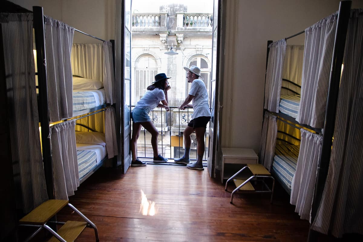 A dormitory room with bunk beds, a small wooden table, and a view of a building through an open door.