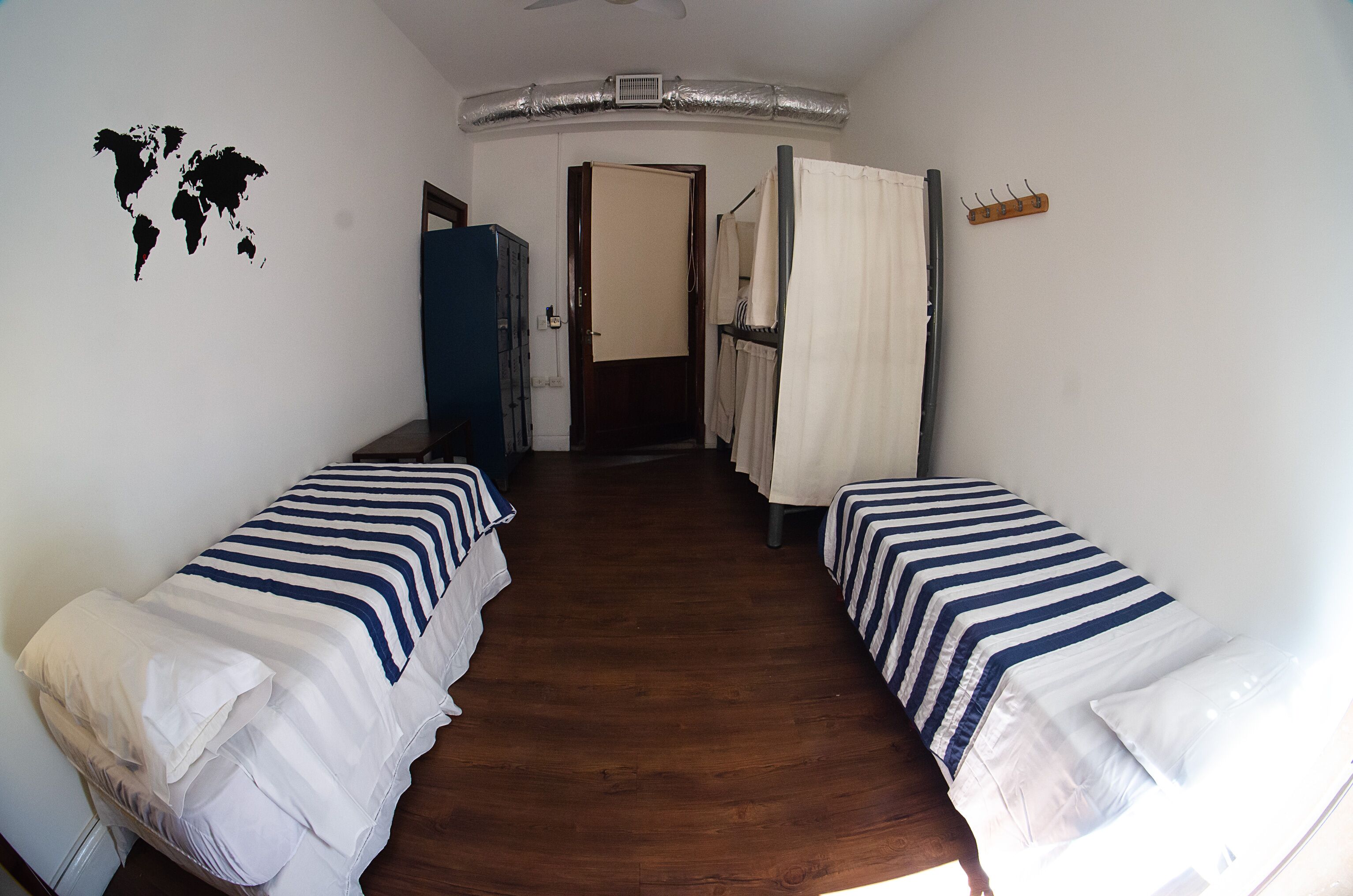 Une chambre avec deux lits simples, une carte du monde accrochée au mur, un plancher en bois et une porte donnant sur une autre pièce.