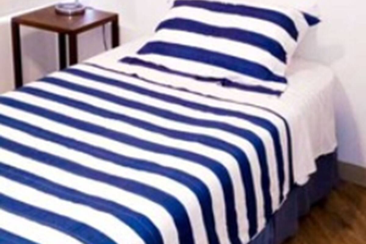 A single bed with a striped bedspread in a room with a wooden floor and a bedside table with a lamp.