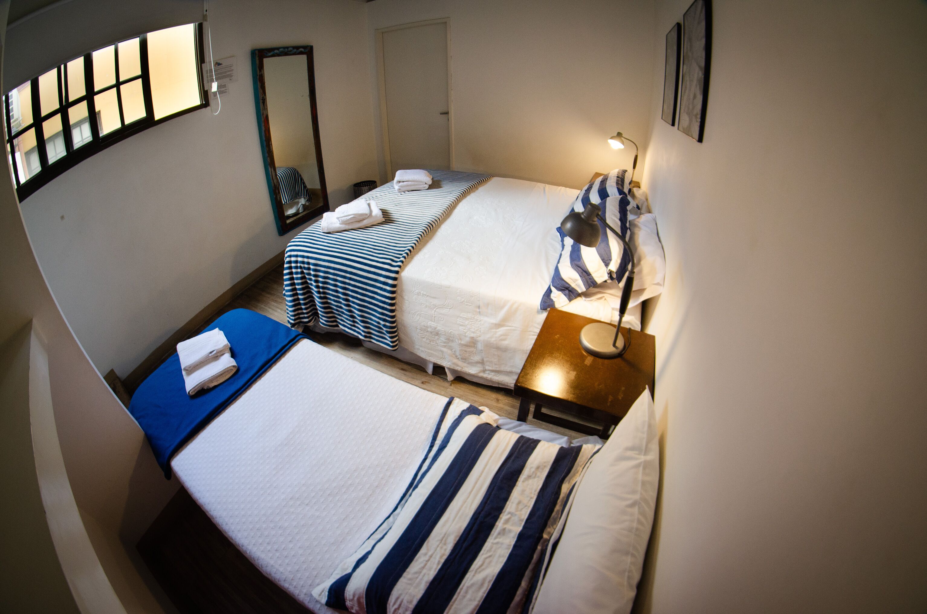 A room with two beds, each with a striped bedspread and pillows, a bedside table with a lamp, and a window with blinds.