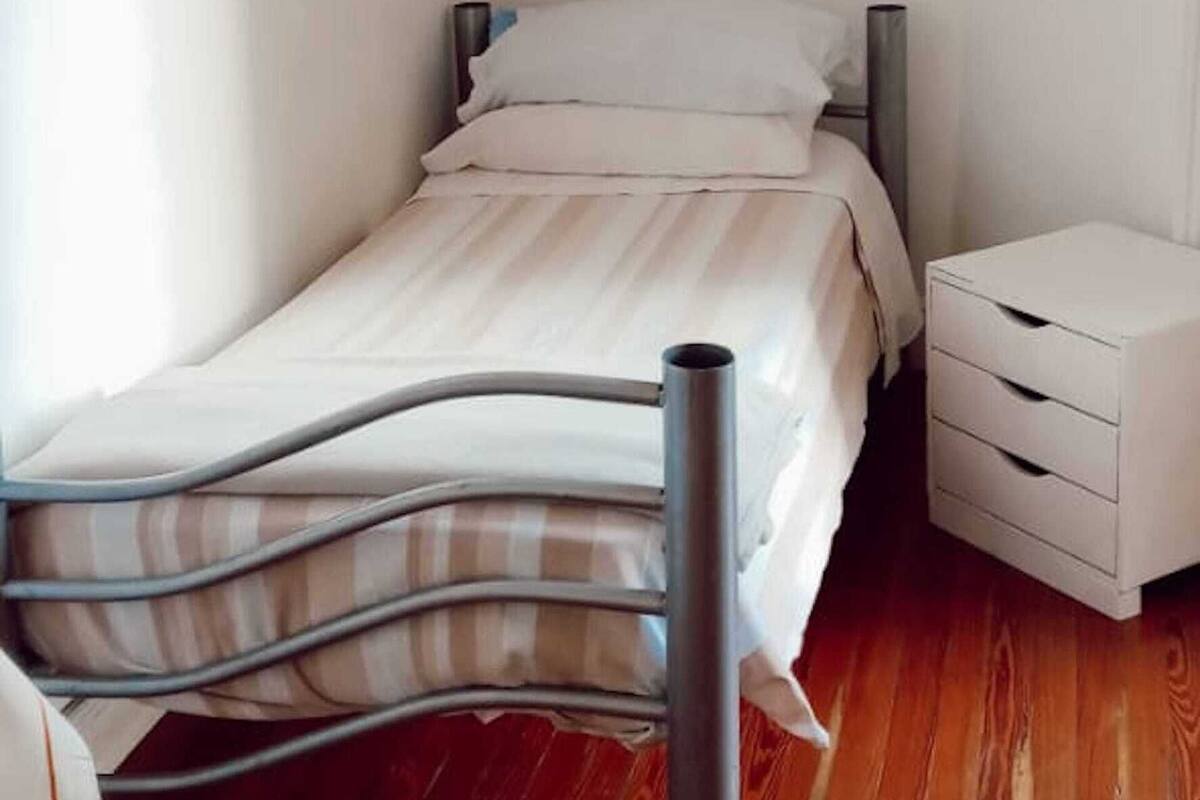 A single bed with a metal frame, white bedding, and a small bedside table with drawers.