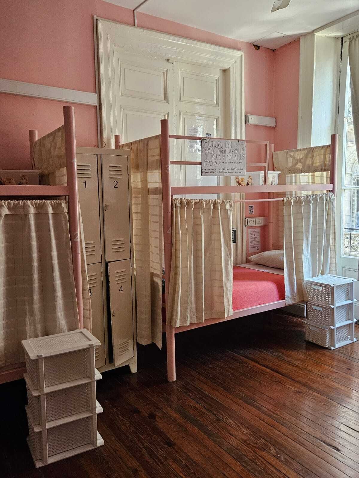 A bunk bed room with a pink wall, wooden flooring, and a window with curtains. There are storage boxes and a sign on the wall.