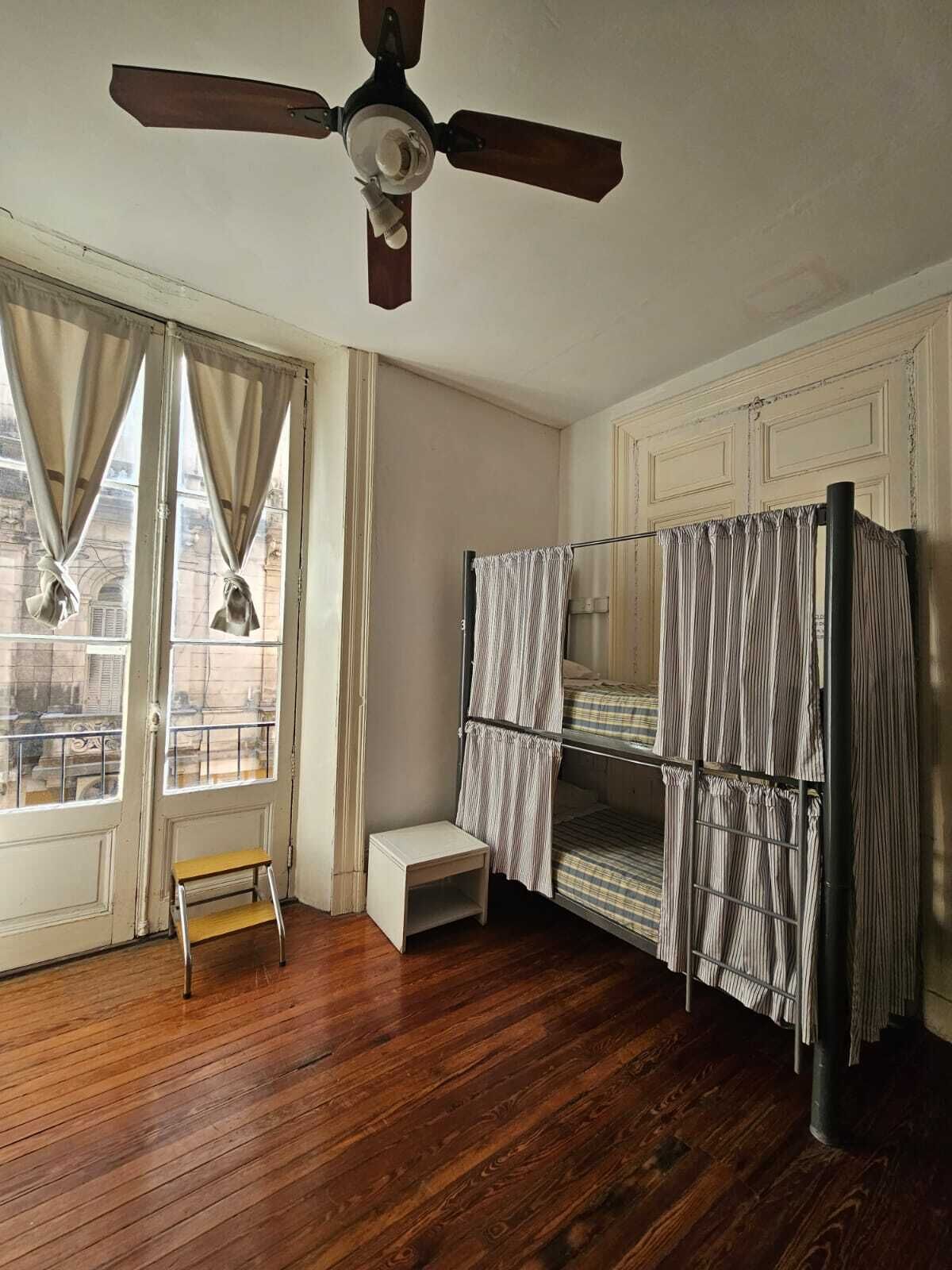A room with a wooden floor, a ceiling fan, a window with curtains, a bunk bed with striped bedding, and a small wooden stool.