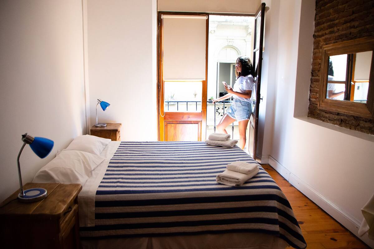 A bedroom with a bed, a nightstand, a lamp, and a window with a view of the outside.