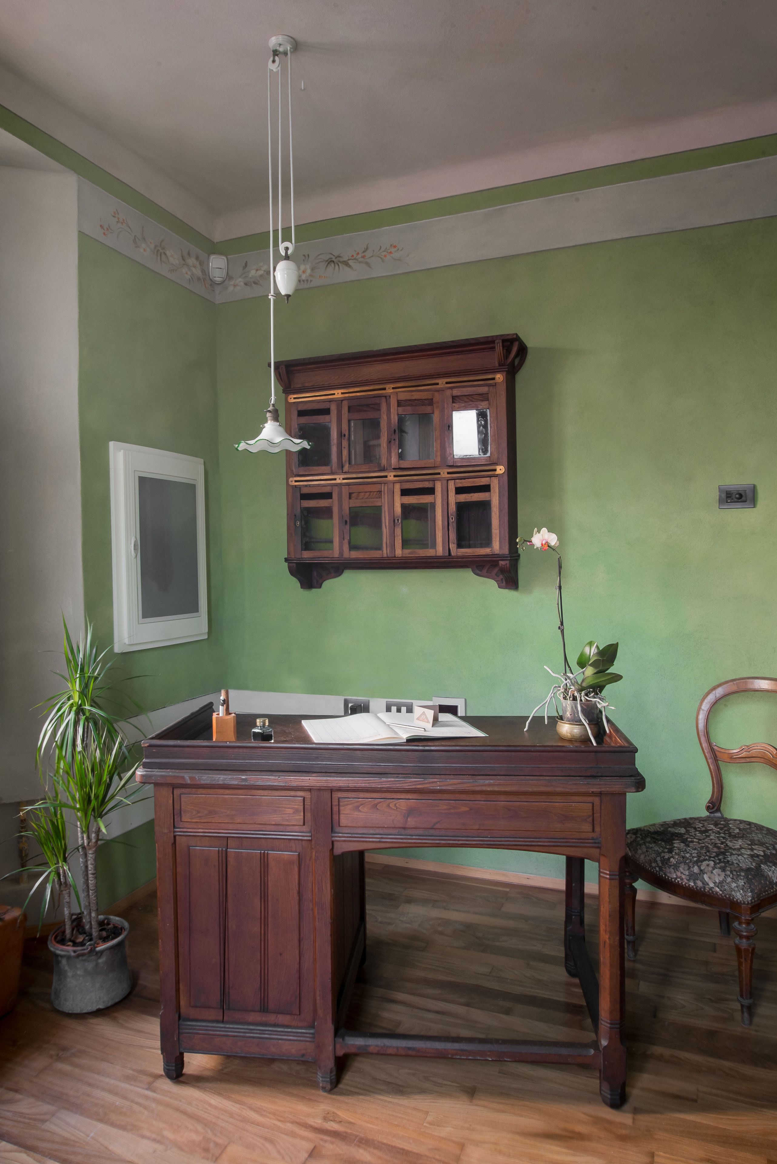 Un bureau en bois avec un évier, une plante en pot et une chaise, dans une pièce aux murs verts et dotée d’une armoire en bois.
