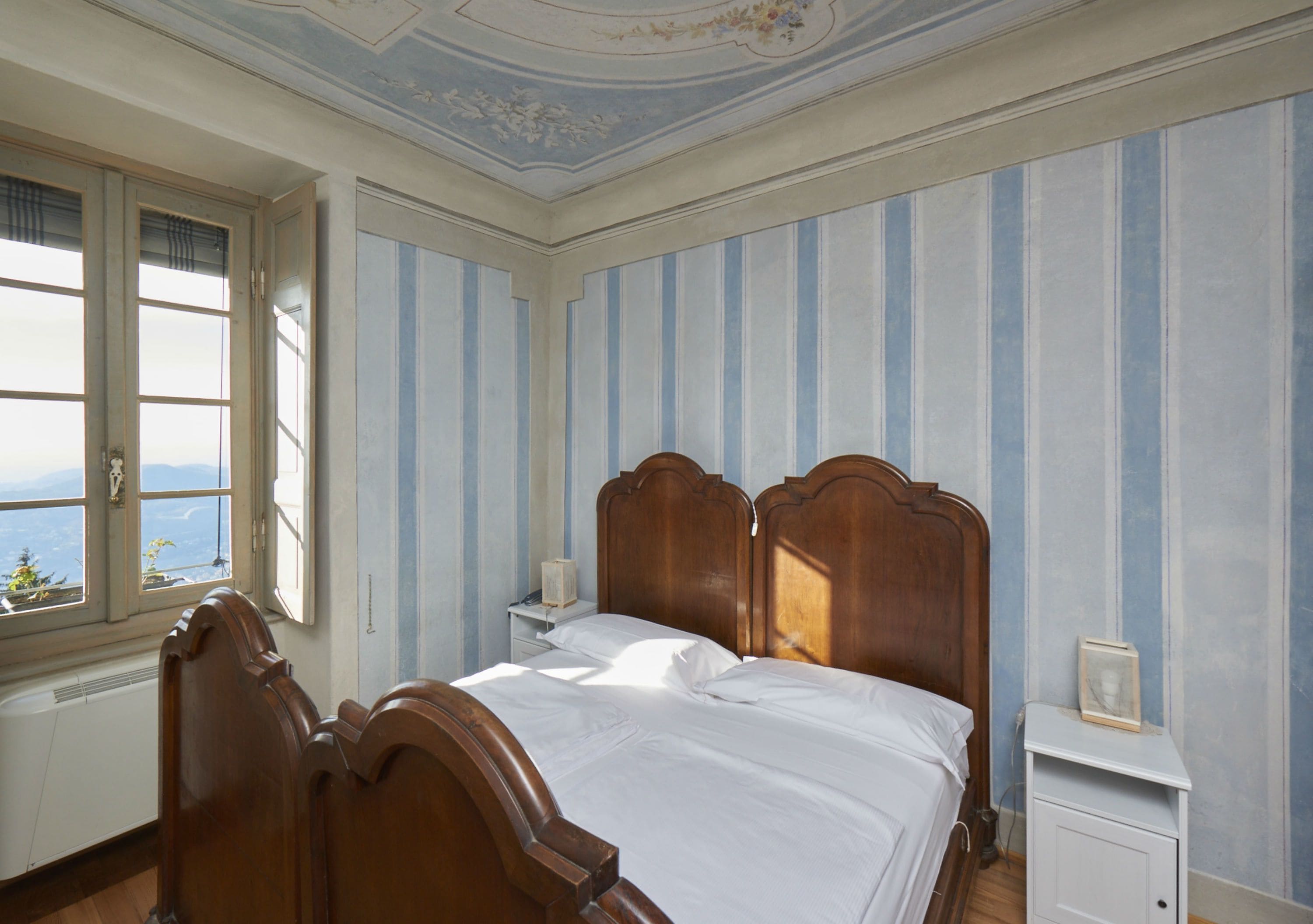 Ein Schlafzimmer mit einem hölzernen Bett, gestreifter Tapete, einem Nachttisch und einem Fenster mit Blick.