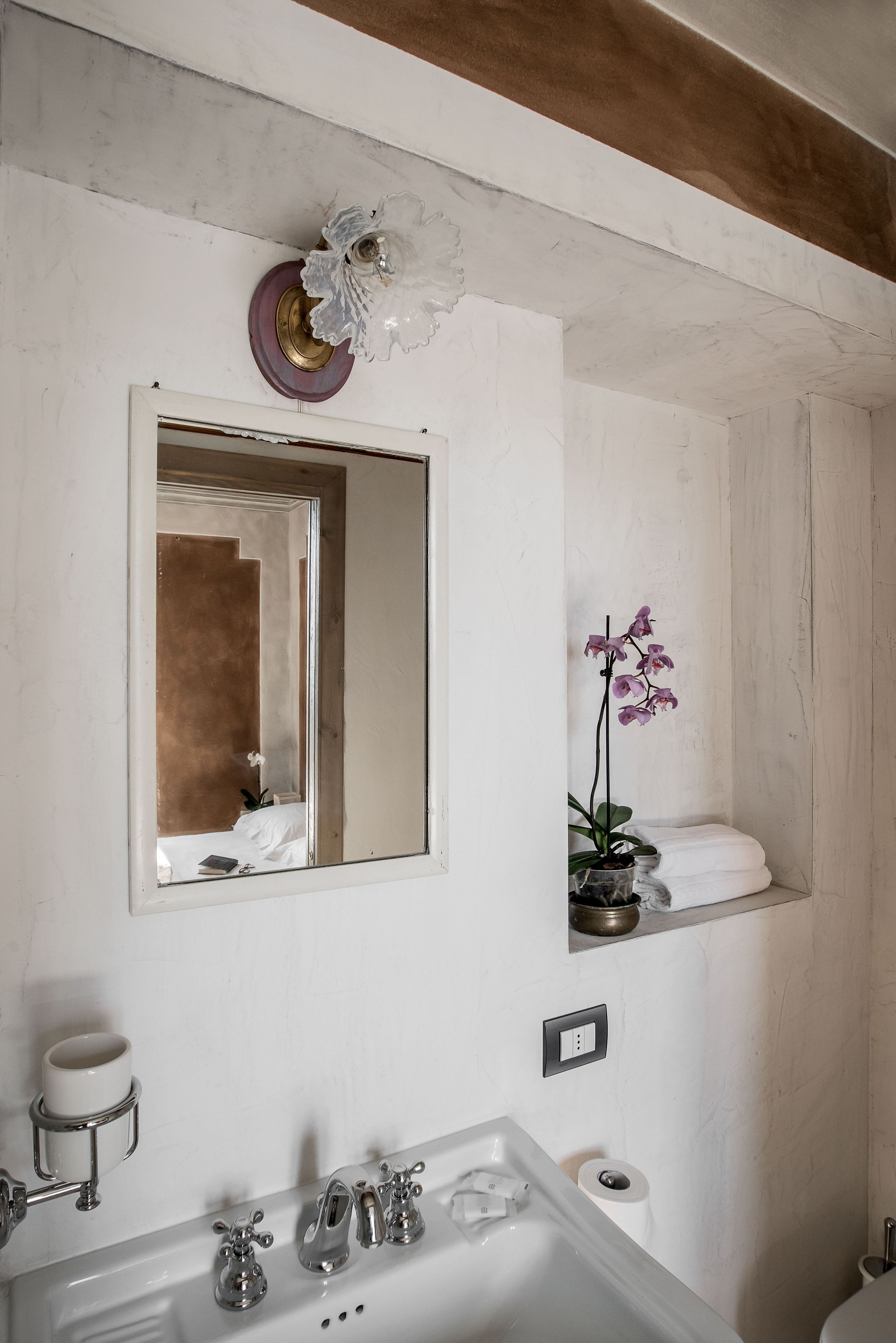 Un bagno con un lavandino bianco, uno specchio, uno scaffale con un'orchidea in vaso e un porta asciugamani.