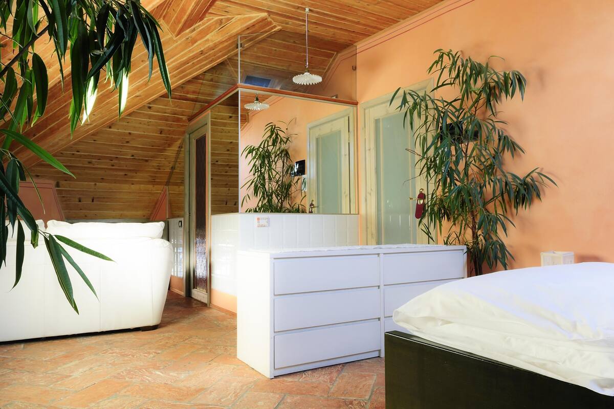 Une chambre avec un lit blanc, une commode blanche et un grand miroir. On y trouve deux plantes en pot, une près du lit et l’autre près de la commode. La pièce est dotée de poutres apparentes en bois au plafond et d’un sol carrelé.