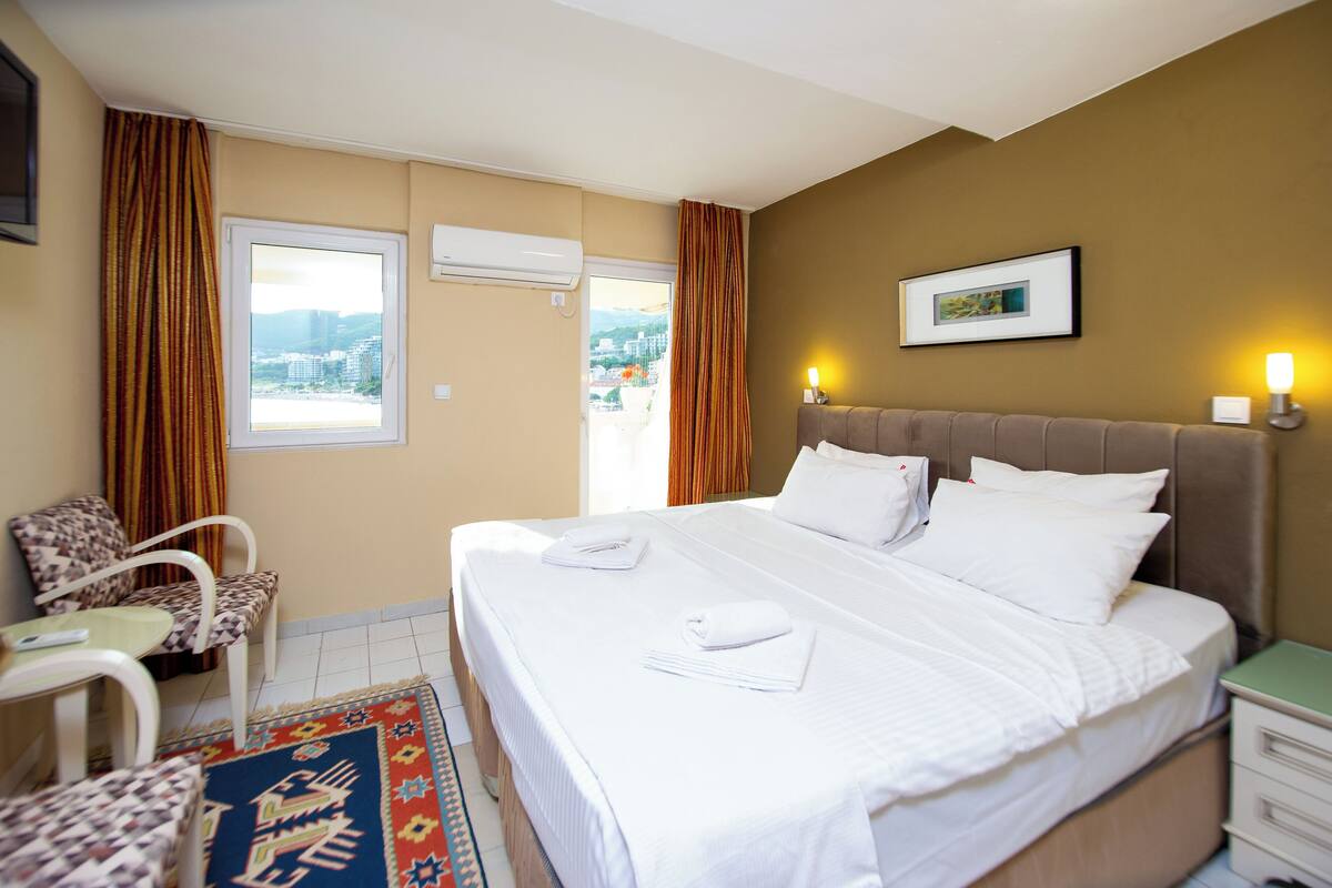 A hotel room with a bed, a chair, a small table, a nightstand, and a window with curtains.