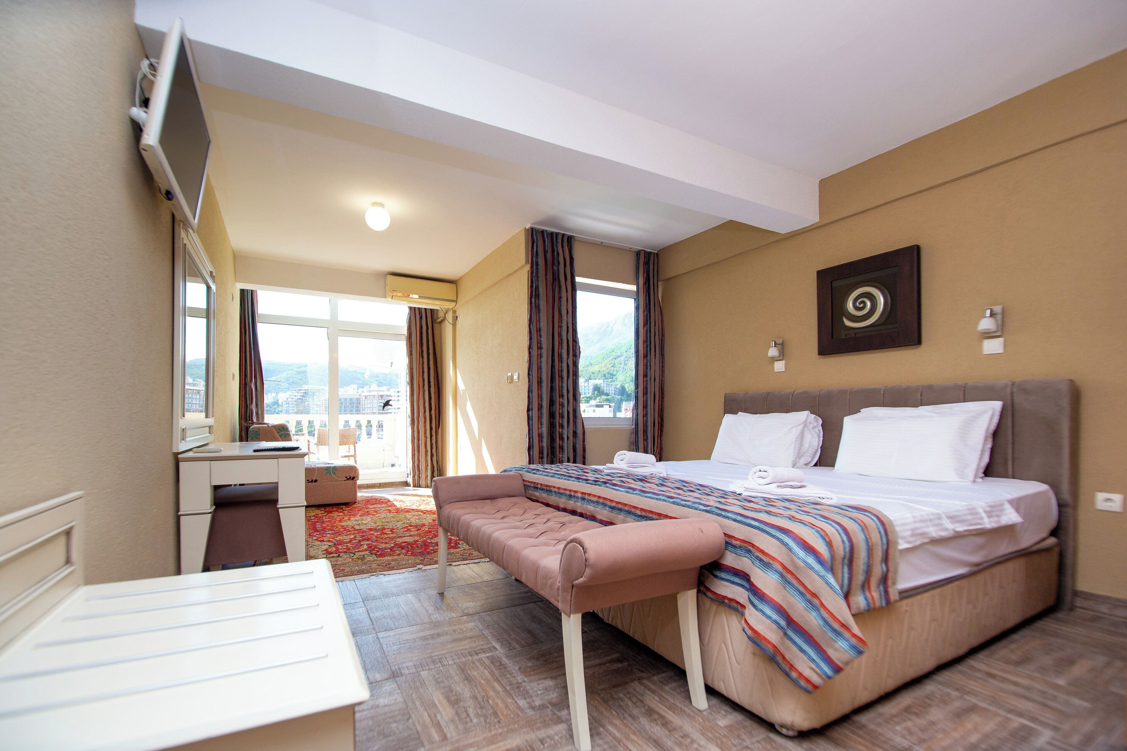 A hotel room with a bed, a desk, a bench, a window with curtains, and a view of the outdoors.