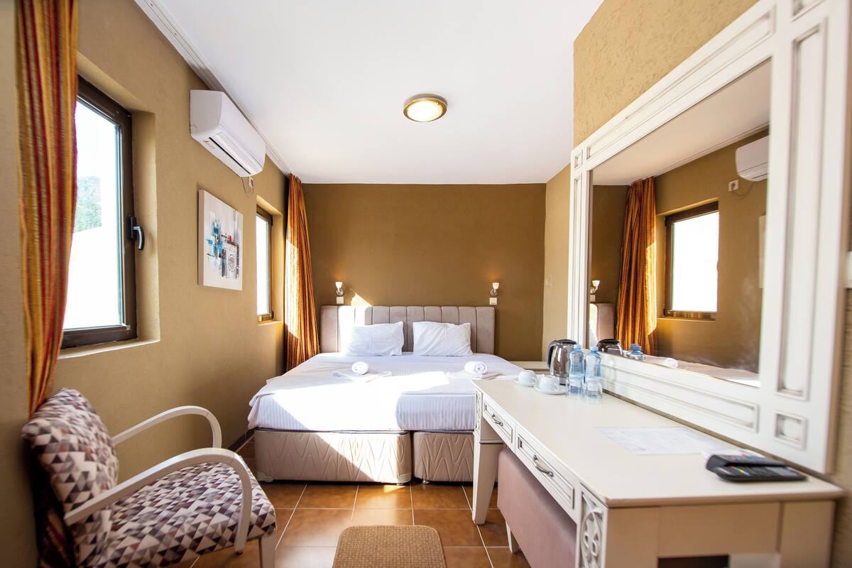 A hotel room with a bed, a desk with a mirror, a chair, and an air conditioning unit.