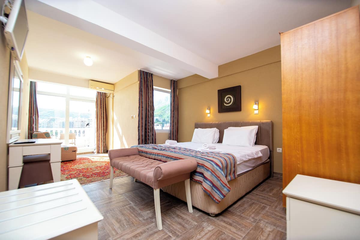 A hotel room with a bed, a desk, a chair, a bench, and a view of the city through large windows.