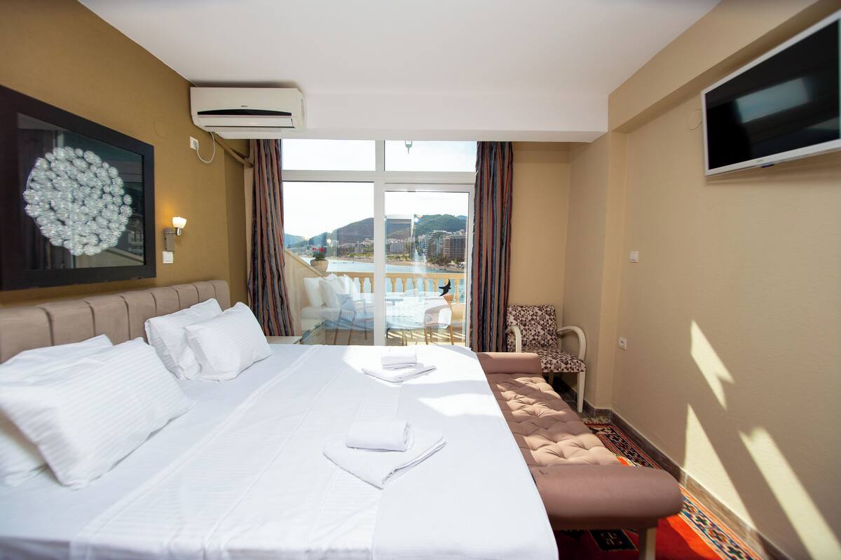 A hotel room with a large bed, a sofa, a TV, and a view of a body of water and buildings.