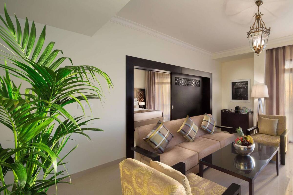Una sala de estar moderna con un sofá, sillones, una mesa de centro y un televisor. Hay una planta grande y un espejo decorativo en la pared.