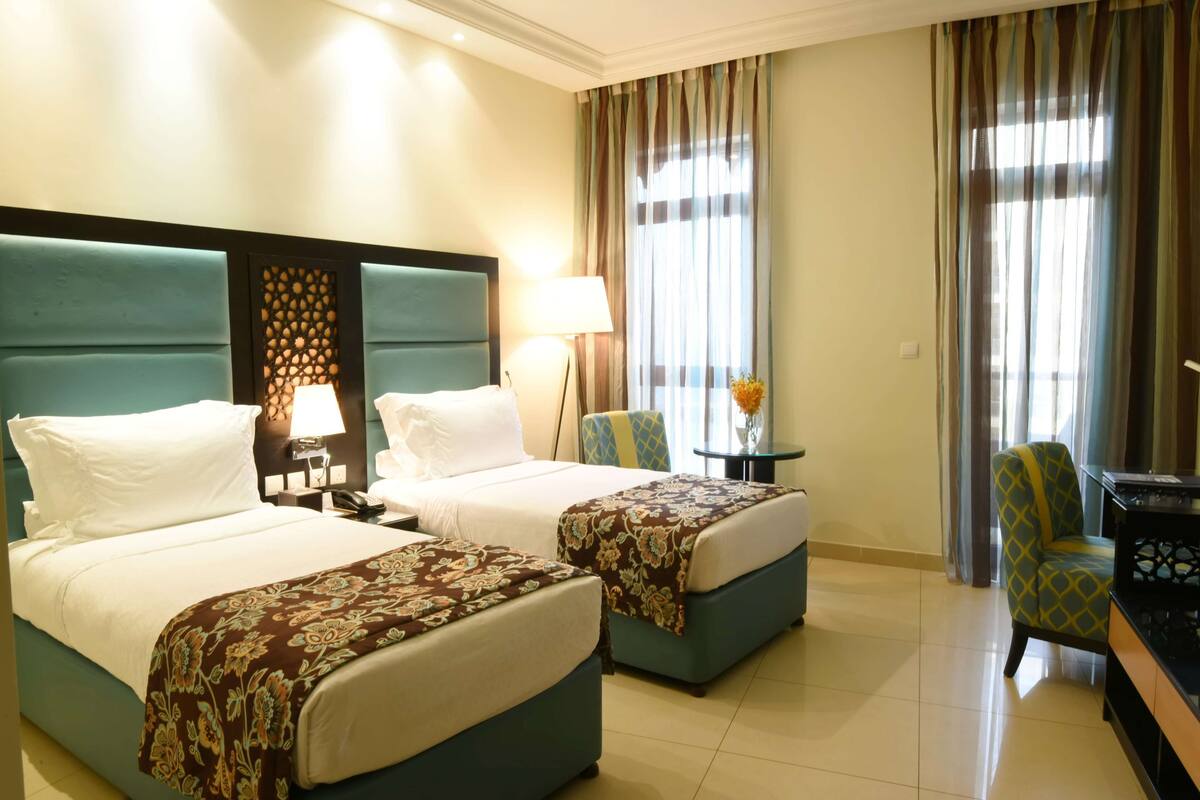 Habitación de hotel con dos camas, una mesita de noche con lámpara, una silla y una pequeña mesa con un florero.