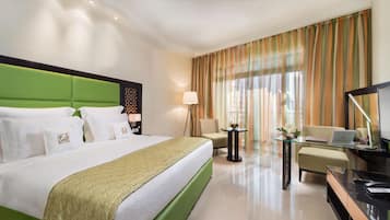 A hotel room with a green upholstered headboard, a large bed with white linens, a flat-screen TV, a small table with a laptop, and a chair.