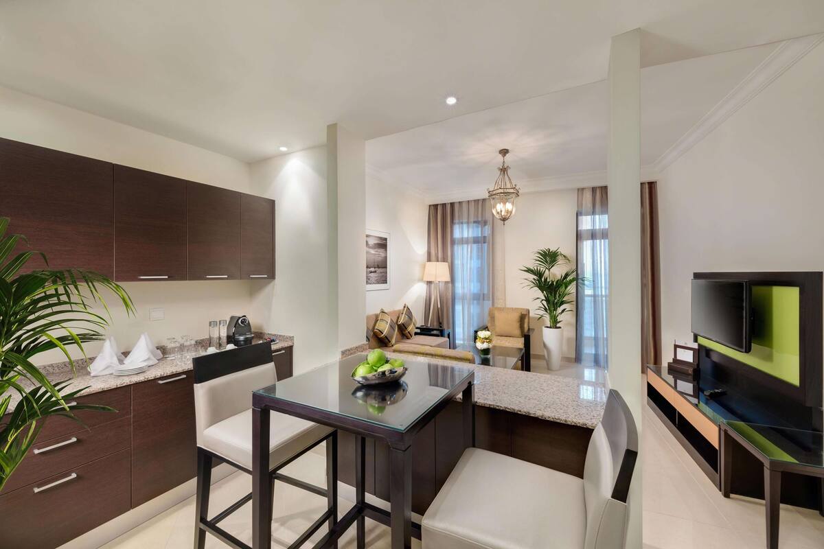 Una cocina moderna con armarios oscuros, un comedor con mesa de vidrio y una sala de estar con sofá y televisor.